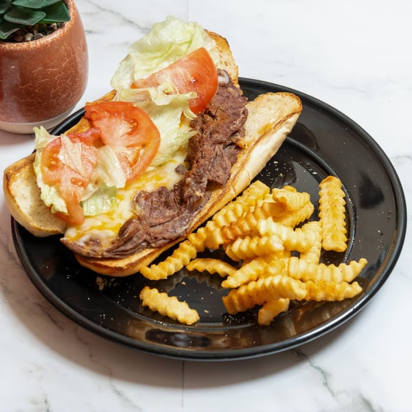 Pete's Pizza cheesesteak with melted cheese, lettuce, tomatoes, and crinkle-cut fries on a black plate.