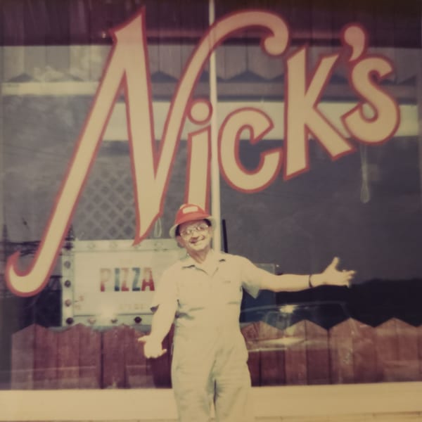 Smiling man in a hat and overalls gesturing in front of a window with "Nick's" and "Pizza" signs.