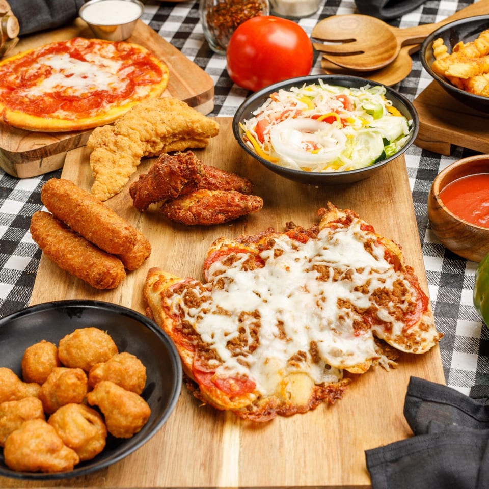 A feast of Pete's Pizza dishes, including pizzas, chicken tenders, wings, and sides on a checkered tablecloth.