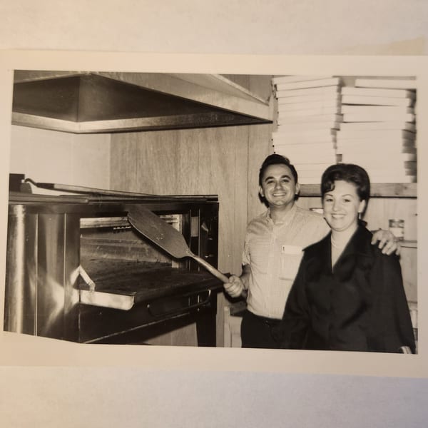 Smiling couple, one holding a pizza peel, standing in front of a pizza oven at Pete's Pizza.