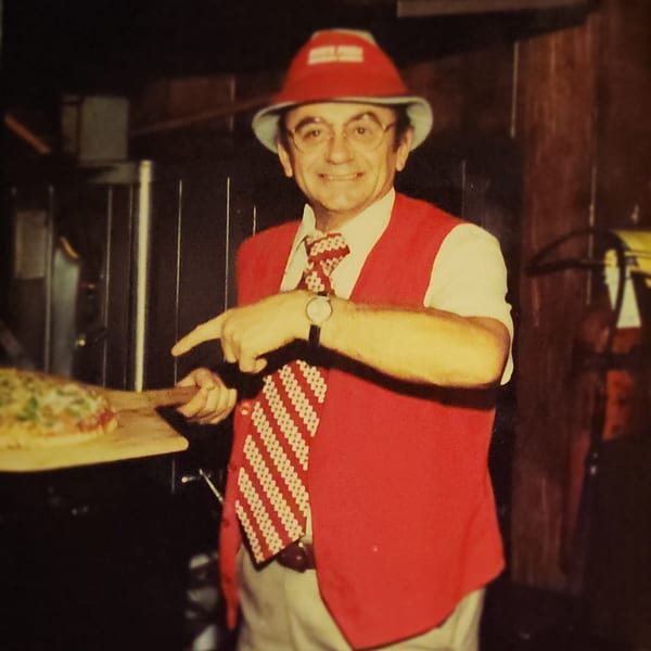 Smiling man in a red vest and hat, holding a pizza on a peel, pointing towards it.