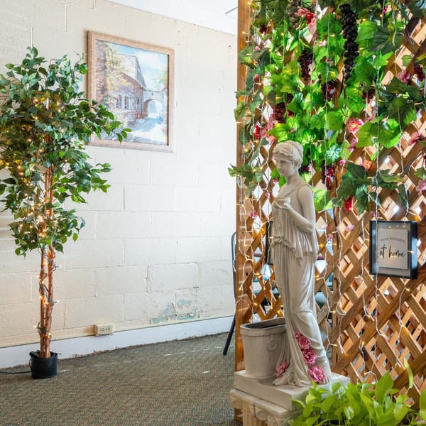 A statue of a woman holding a cup stands next to a wooden trellis adorned with grapevines and lights.