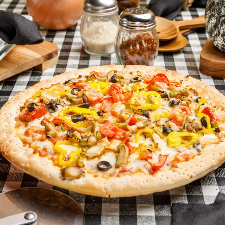 Pete's Pizza with colorful toppings, including yellow peppers, tomatoes, olives, and mushrooms on a checkered tablecloth.