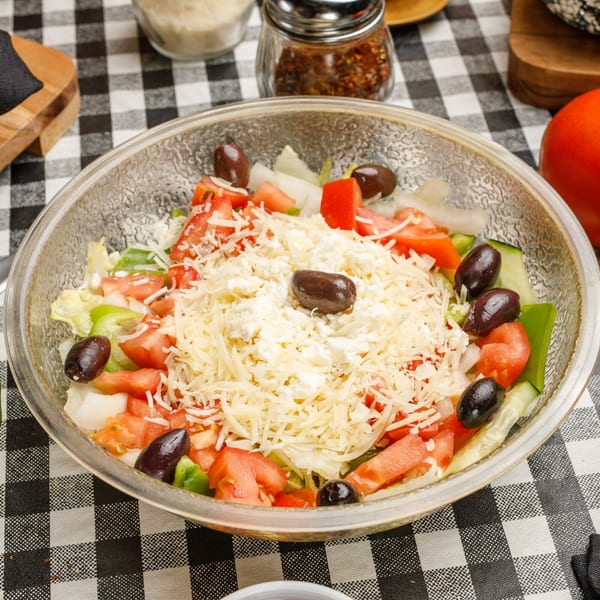 A fresh Pete's Pizza salad with tomatoes, olives, and cheese in a clear bowl on a checkered tablecloth.