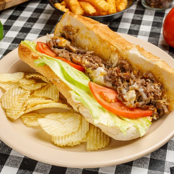 Cheesesteak sandwich with lettuce, tomato, and a side of ripple chips on a black and white checkered tablecloth.